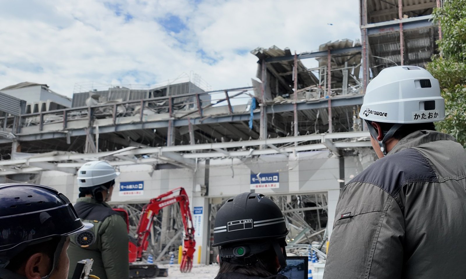 爆発で外壁や上層部が大きく損壊したイオンモール熊本を前に、ヘルメット姿の災害支援関係者が機材を確認する様子