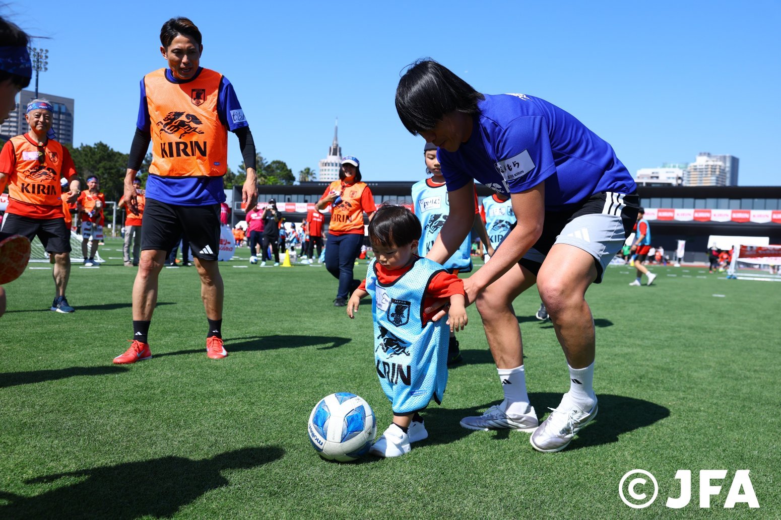 芝生のグラウンドで大人に支えられながらサッカーボールを蹴る幼児