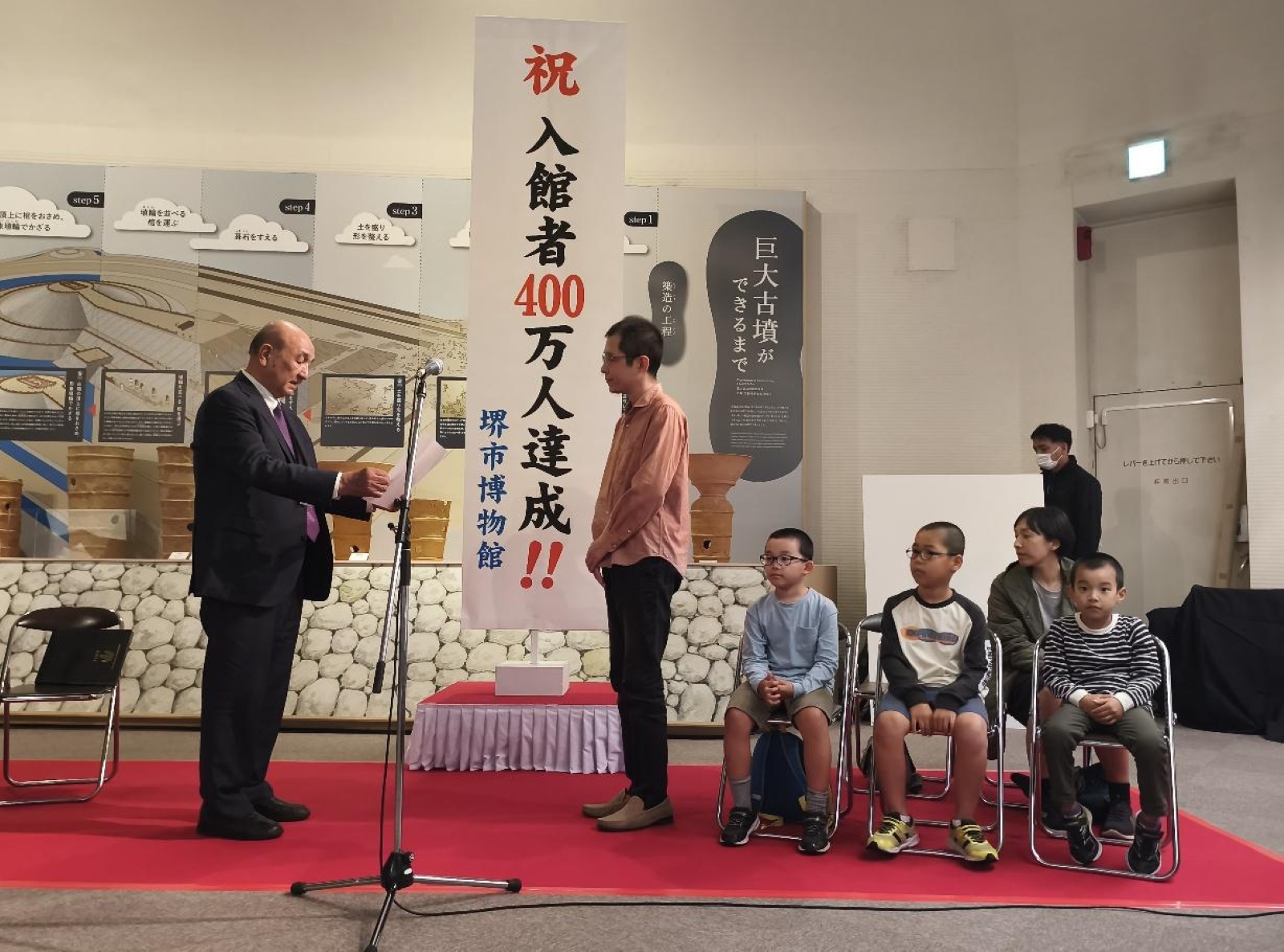 堺市博物館の累計入館者400万人達成記念セレモニーで、浜崎さんに記念状を贈呈する博物館長。そばには浜崎さんの家族が座っている
