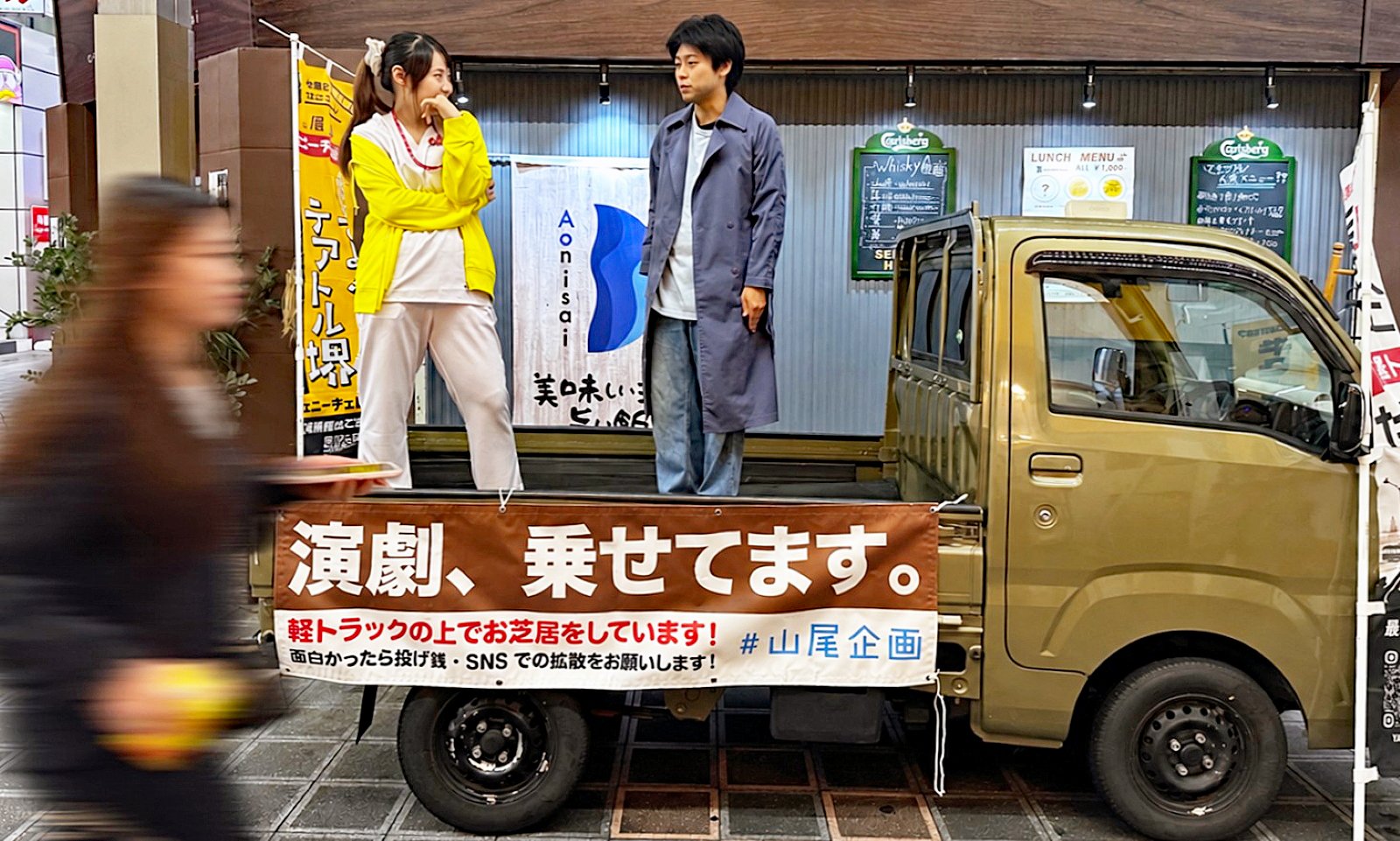 堺東の商店街で軽トラックの荷台に立って演じる山尾匠さんと共演者