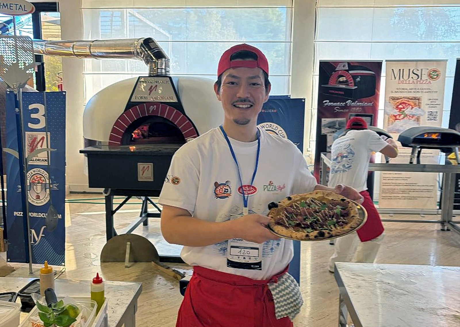 イタリアで開催されたピッツァ世界大会の会場で、焼き上げたピッツァを手に笑顔を見せる洲鎌健人さん。背後にはピザ窯が並ぶ